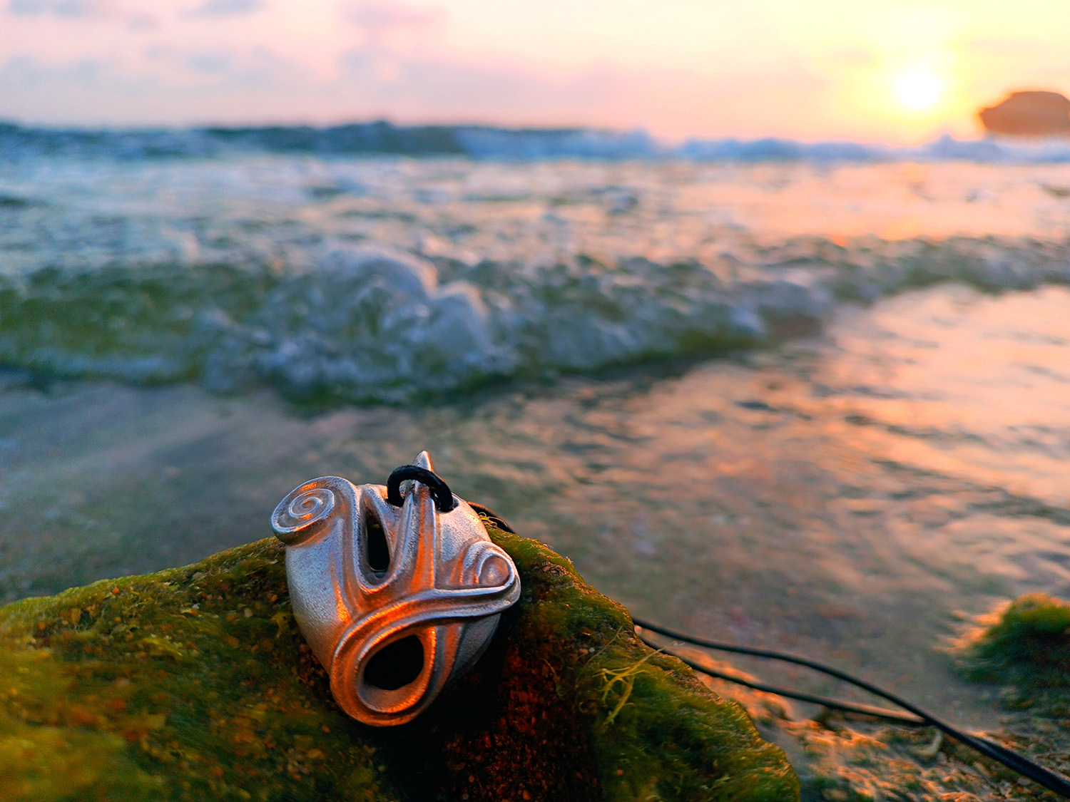 Ein silberner Halskettenanhänger mit einem komplizierten Design liegt auf moosbedeckten Felsen am Ufer. Im Hintergrund plätschern sanfte Wellen an den Strand, während ein leuchtender Sonnenuntergang warme Farbtöne über Himmel und Wasser wirft.
